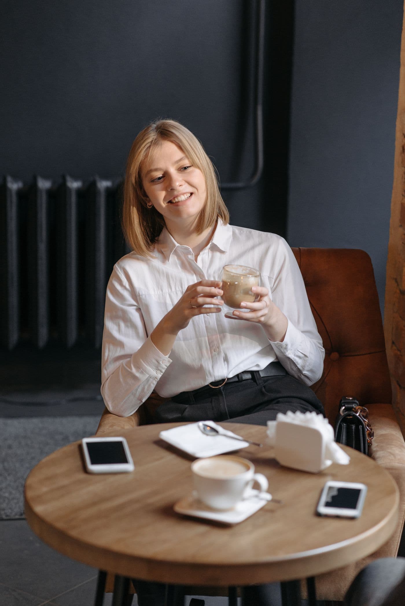 A smiling blonde woman in a white shirt holding a coffee at a café table